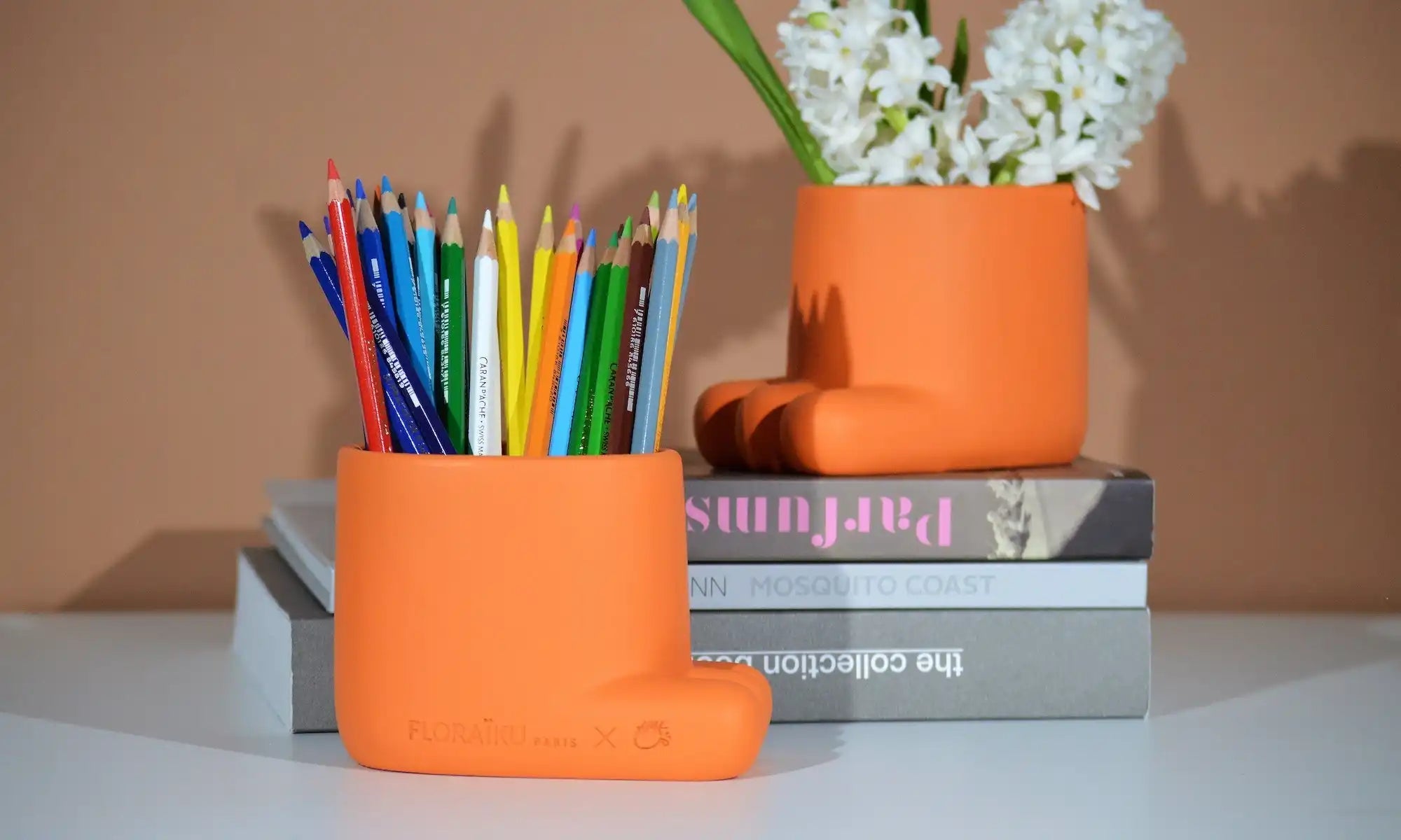 Orange ceramic or clay pot holding colorful pencils, with a matching pot containing white flowers beside it.